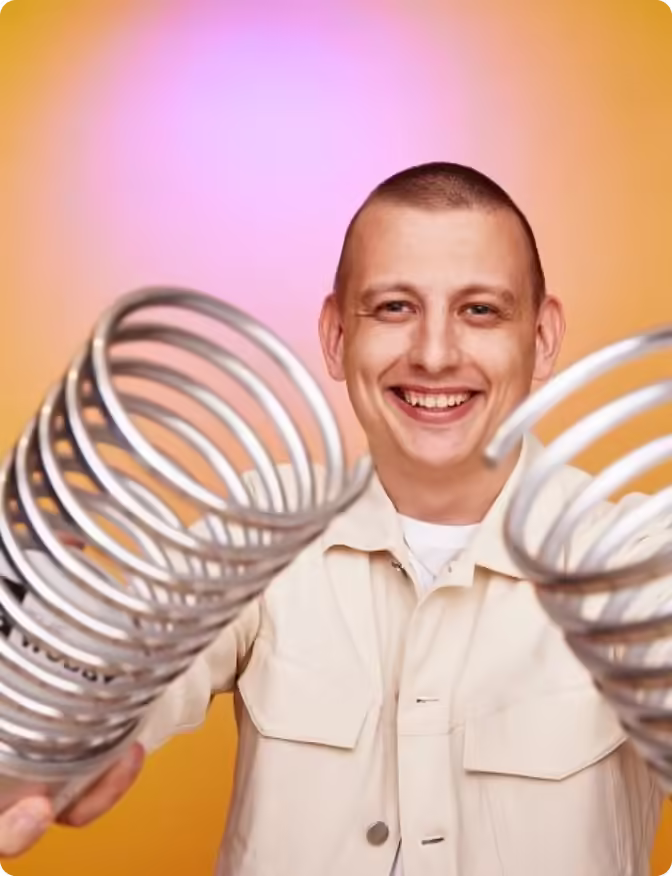 Smiling man holding two large metal springs toward the camera over an orange and pink background.