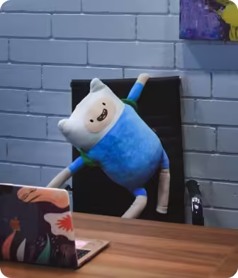 Plush toy wearing blue sits on a black chair behind a desk with a laptop showing colorful stickers.