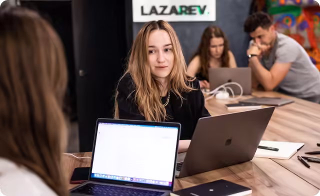 Young people working on laptops around a table in a modern office setting with Lazarev sign in background.