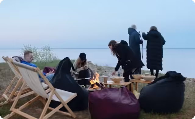 Group of people gathered around a campfire on a beach in the evening with chairs and bean bags.