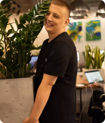 Smiling person leaning on a planter with green leafy plants in a cozy office space.