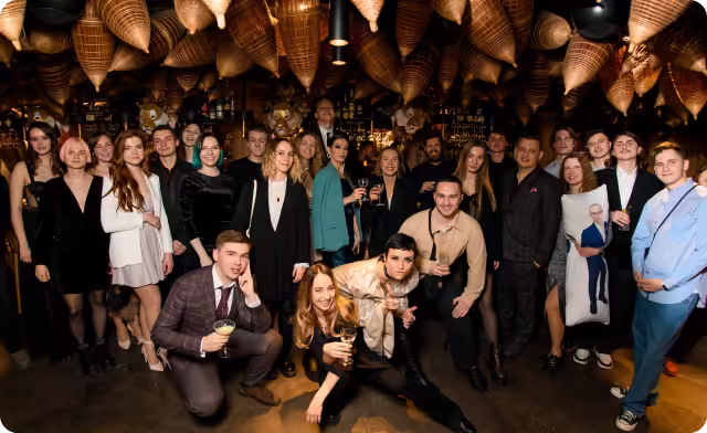 Group of formally dressed young adults posing and holding drinks under hanging wicker lamps in a dim bar.