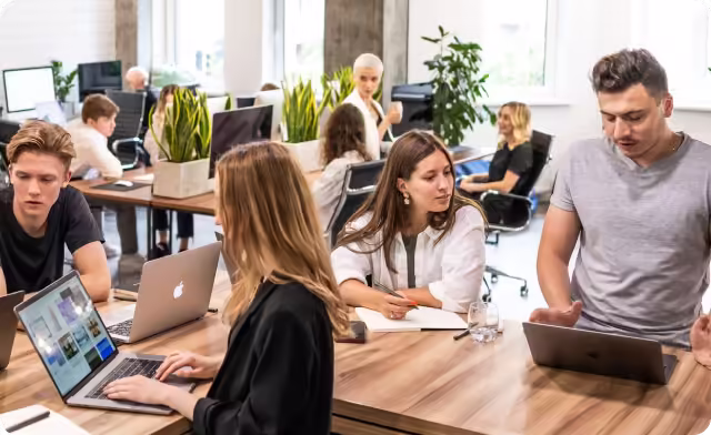 Young professionals collaborating at a large wooden desk in a bright modern office with laptops and notebooks.