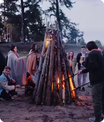 Group of people around a tall campfire in a forested area at dusk, some roasting food on sticks.