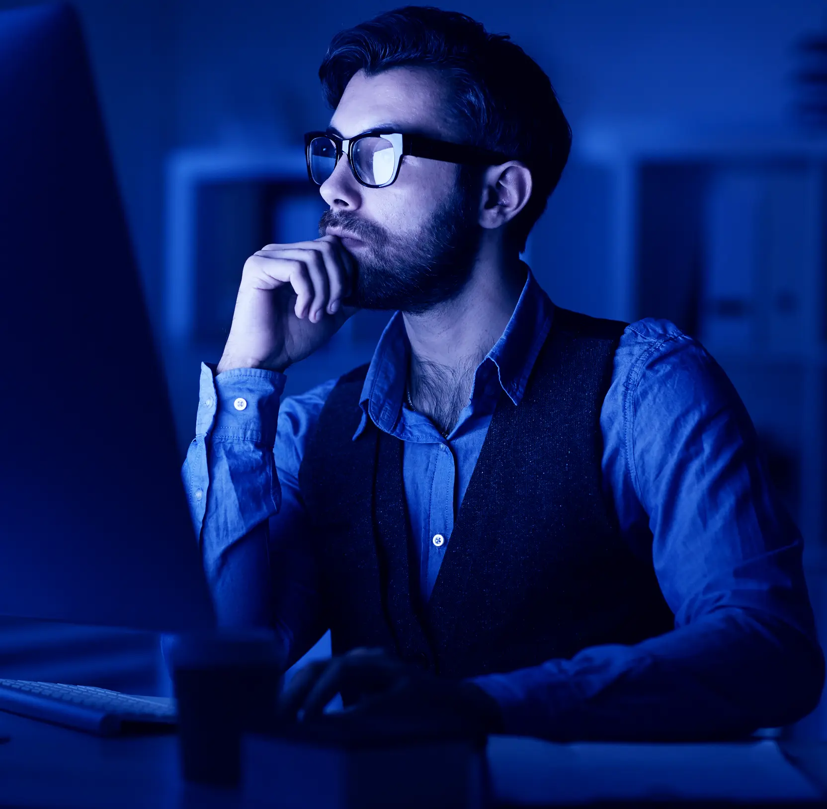Man in glasses reviewing data on a monitor, lit by blue screen light