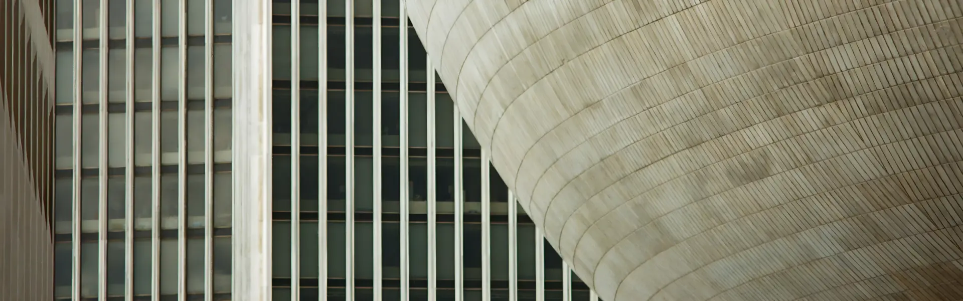 Architectural close-up showing the contrast between a modern high-rise building with vertical glass and steel window panels in the background, and a large, curved, ribbed concrete structure in the foreground.