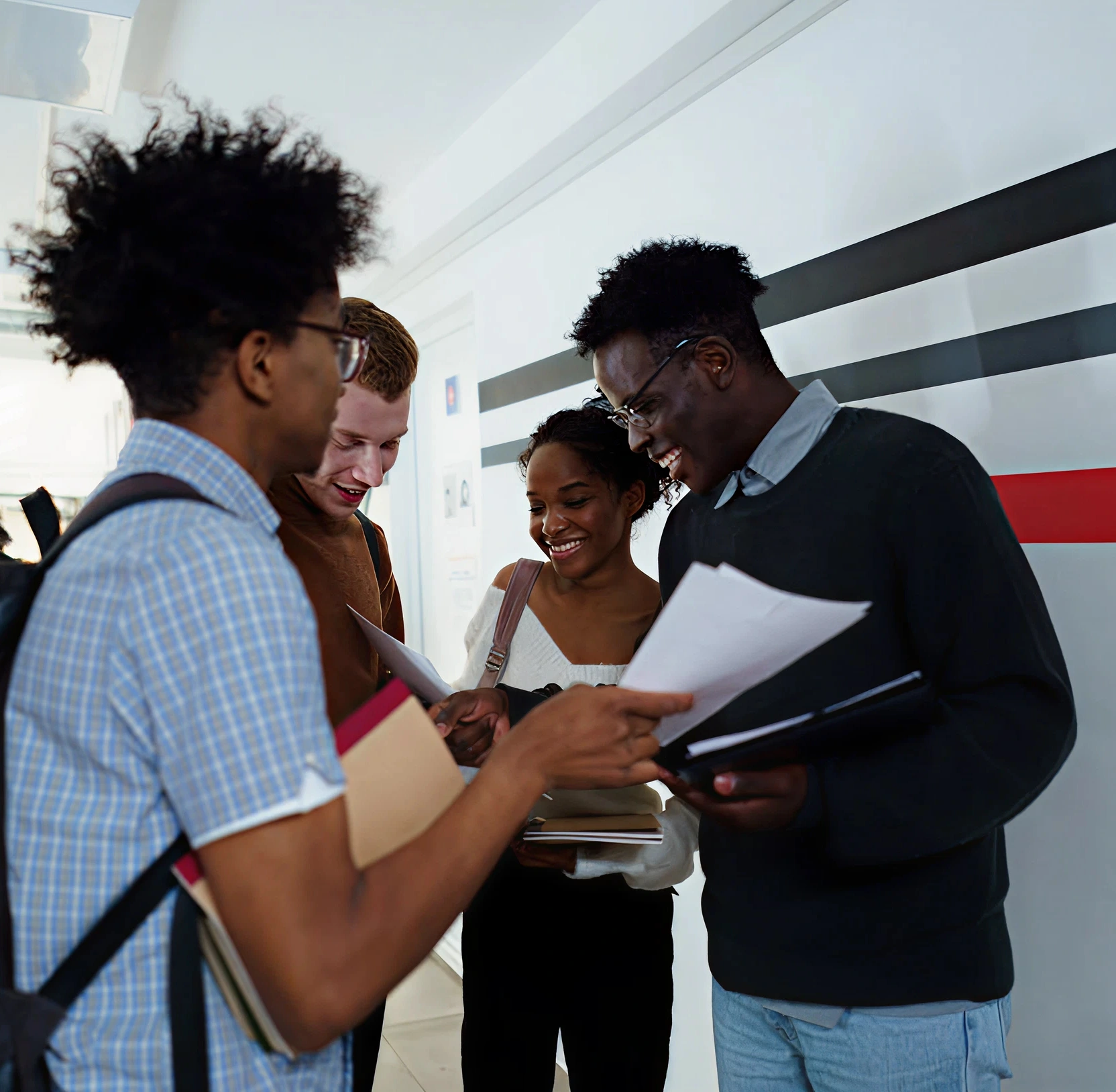 Four international students stand together in a bright campus hallway, smiling as they look over printed documents.