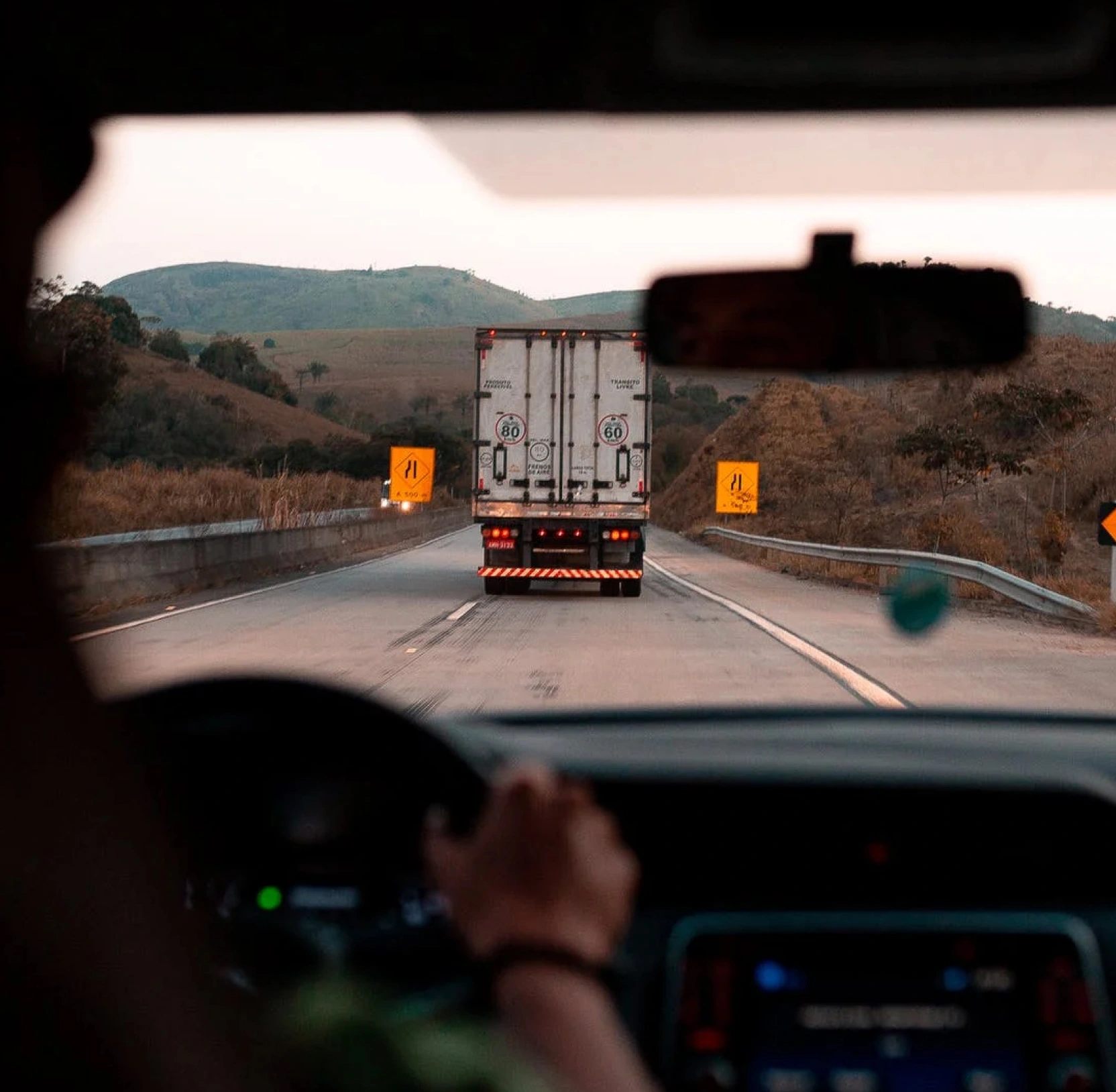 Driver's point of view through a windshield while following a cargo truck along a highway through rolling hills at dusk.