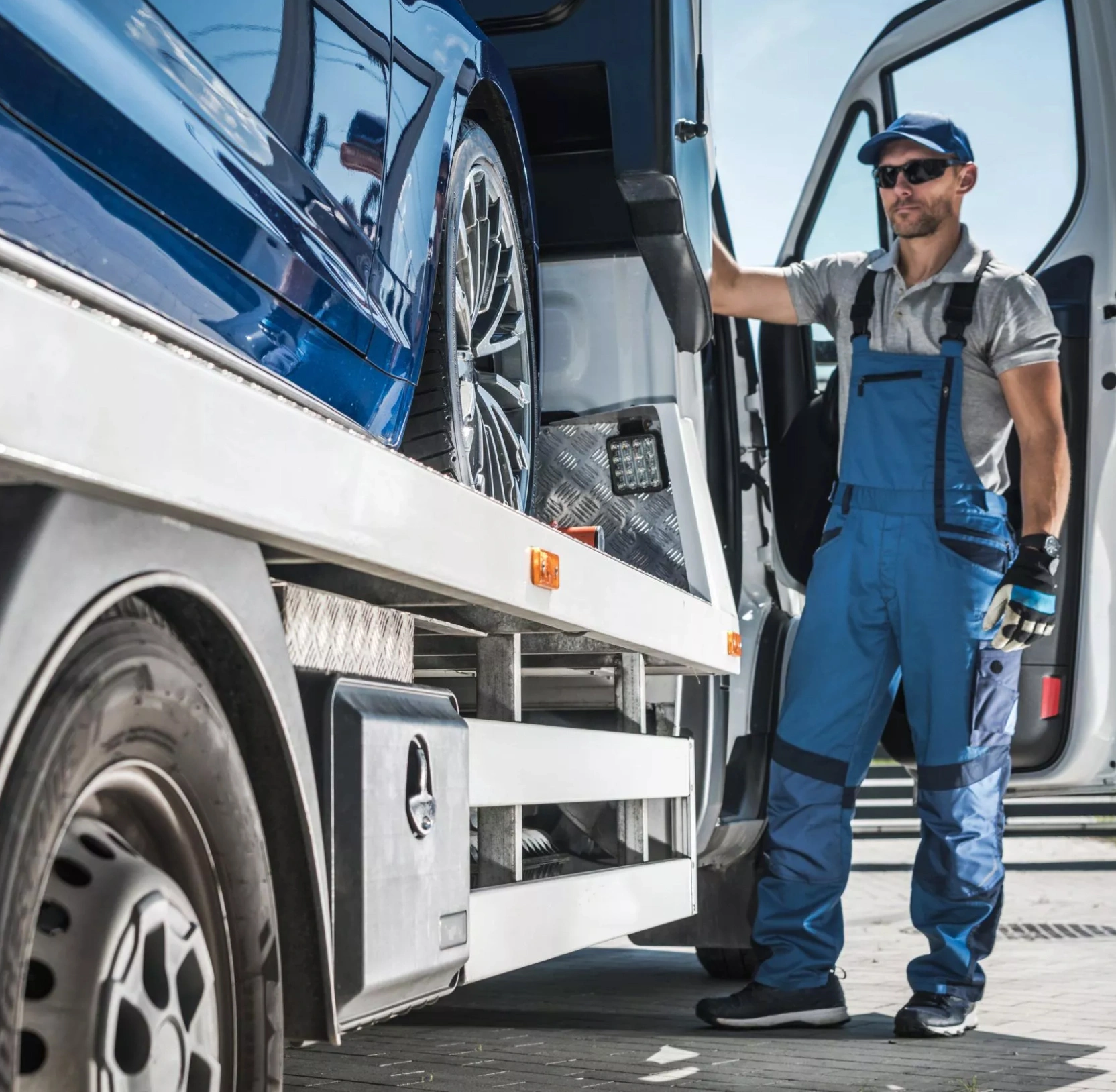 Auto-transport worker in blue coveralls standing beside a flatbed carrier loaded with a blue sports car, ready for shipping.